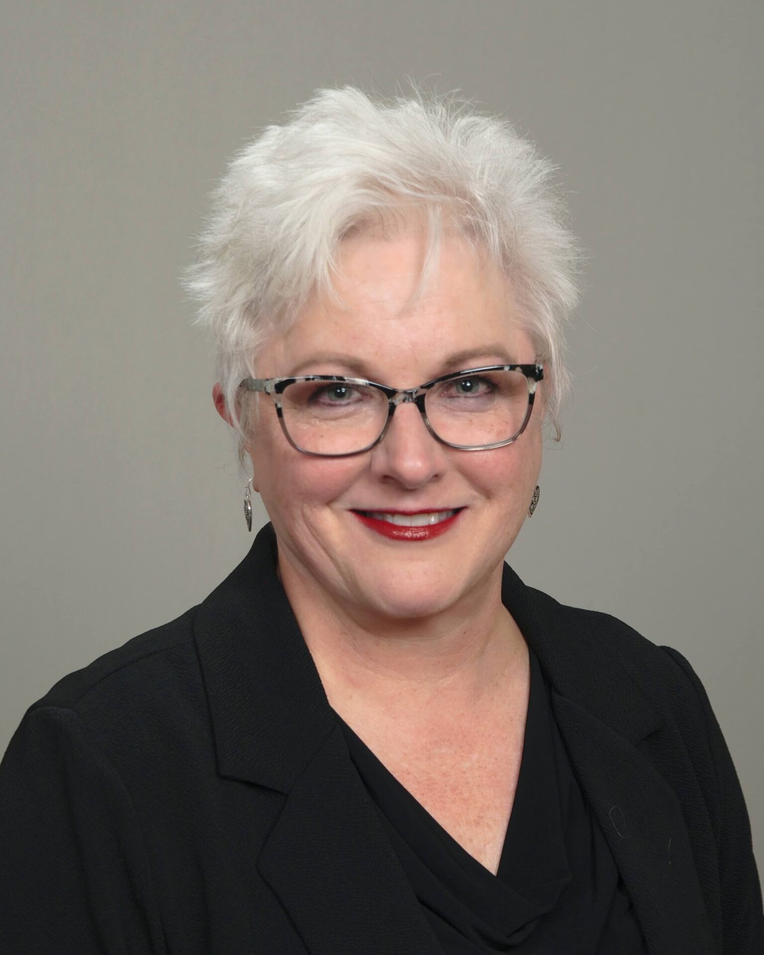 A woman with white hair and glasses in front of a gray background.